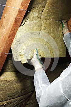 A man in a protective overalls puts mineral wool between the beam of the roof of the house for his warming from the cold