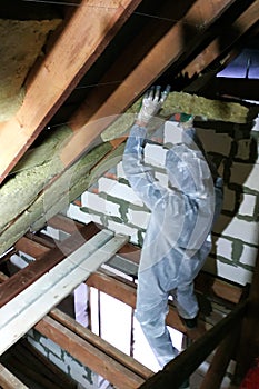 A man in a protective overalls puts mineral wool between the beam of the roof of the house for his warming from the cold