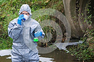 A man in a protective suit, adjusts the mask on the background of drainpipes, looking forward, holding a tube of liquid