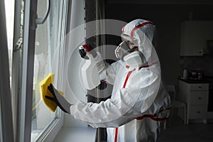 A man in a protective suit cleans the window with a chemical agent, protecting the house from coronavirus