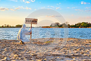 Man in protective suit and gloves sets warning sign with inscription closed along the sand beach