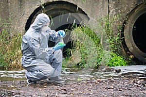 A man in a protective suit and mask, against the drainpipes, holding a tube of green liquid