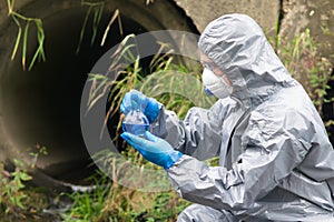 A man in a protective suit and mask holds in his hands a flask with a blue liquid on the background of a large sewage pipe