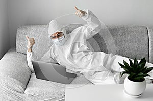 Man in protective suit and medical mask is lying on a sofa in his living room with a laptop and showing two thumbs up