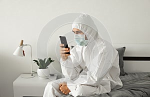 Man in protective white suit and medical mask is using a phone at his home sitting on a bed because of coronavirus