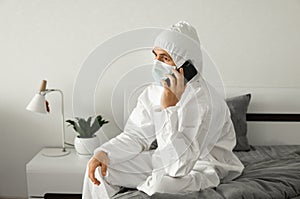 Man in protective white suit and medical mask is using a phone at his home sitting on a bed because of coronavirus