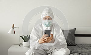 Man in protective white suit and medical mask is using a phone at his home sitting on a bed because of coronavirus