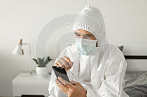 Man in protective white suit and medical mask is using a phone at his home sitting on a bed because of coronavirus