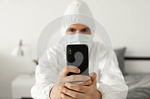 Man in protective white suit and medical mask is using a phone at his home sitting on a bed because of coronavirus