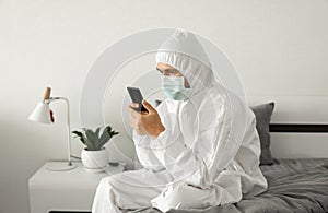 Man in protective white suit and medical mask is using a phone at his home sitting on a bed because of coronavirus