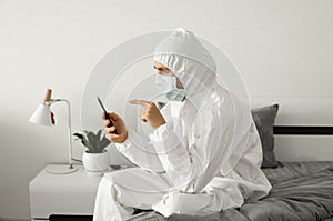 Man in protective white suit and medical mask is using a phone at his home sitting on a bed because of coronavirus