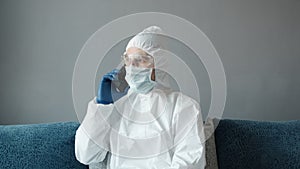 Man in protective white suit and medical mask is using a phone at his home sitting on a bed because of coronavirus