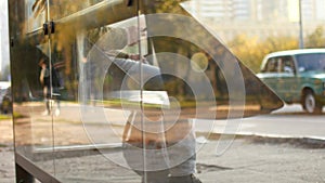 A man at a public transport stop is waiting for a bus. A man uses mobile Internet while waiting for transport