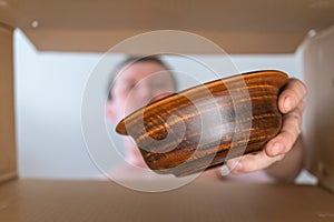 A man pulls a brown bowl out of a cardboard moving box. Brown handmade bowl made of fired clay. Bottom view. Inside view. Close-up