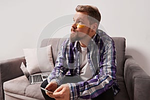 Man in purple shirt with eye patches sitting on the couch with smartphone. Self care routine