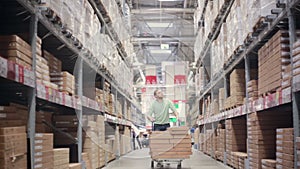 A man is pushing a trolley full of boxes on it between shelves with cardboard boxes in a storage warehouse