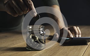 A man put coins in a glass jar and took notes to save money on a wooden table