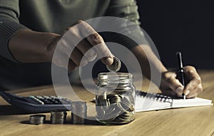 A man put coins in a glass jar and took notes to save money on a wooden table