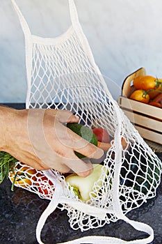 Man puts avocado to the eco shopping bag with fresh vegetables - tomatoes, purple potatoes, eggplants, carrots, sweet