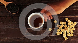 A man puts a cup of tea on the table, on him located brown sugar cubes and a spoon with tea leaves on brown wooden table