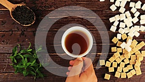 A man puts a cup of tea on the table, on him located brown and white sugar cubes, spoon with tea leaves, mint leaves on