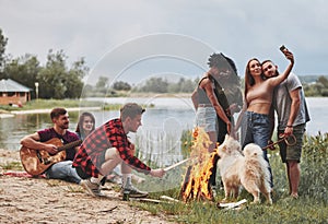 Man puts wood in fire. Group of people have picnic on the beach. Friends have fun at weekend time