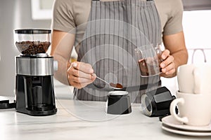 Man putting ground coffee beans into moka pot at table in kitchen, closeup