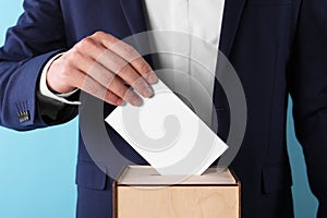 Man putting his vote into ballot box on light blue background, closeup