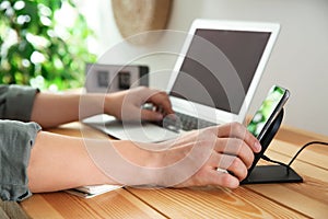 Man putting mobile phone onto wireless charger at table, closeup. Modern workplace accessory