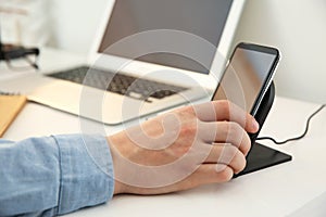 Man putting mobile phone onto wireless charger at table, closeup. Modern workplace accessory