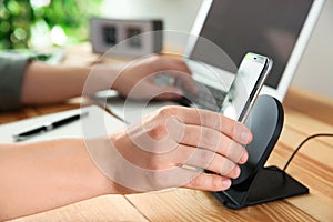 Man putting mobile phone onto wireless charger at table, closeup. Modern workplace accessory