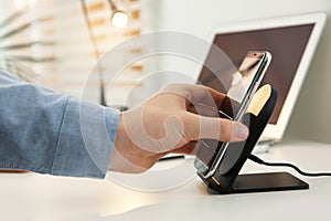 Man putting mobile phone onto wireless charger at white table, closeup. Modern workplace accessory