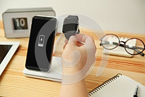 Man putting smartwatch onto wireless charger at wooden table, closeup. Modern workplace accessory