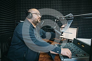 Man radio host or representer or journalist reads news from paper list in hand to studio microphone sitting at mixing sound panel