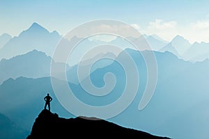 Man reaching summit after climbing and hiking enjoying freedom and looking towards mountains silhouettes panorama during