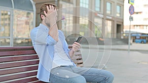 Man Reacting to Loss on Smartphone while Sitting Outdoor on Bench