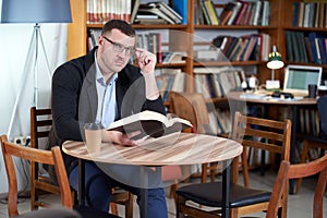 Man reading book and drinking coffee in a library cafe with many books on a background