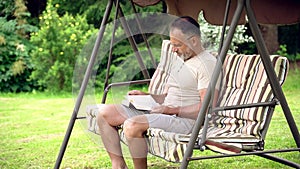 Man reading a book sitting on a garden swing in a green summer garden outdoors