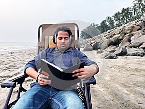 Man reading a book while wearing headphone relaxing on a beach chair with tropical scenery in the background