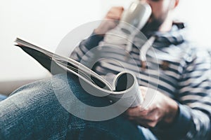 Man reading magazine or newspaper and drinking coffee while relaxing on sofa