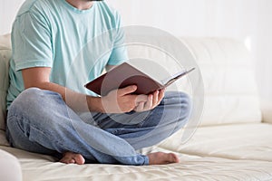 Man reading old hardback note book holding in his hands, on the white couch. Domestic lifestyle. Education concept.