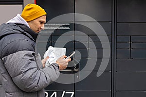 Man receiving parcel from automatic post box using smartphone outdoors. Modern delivery