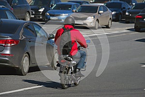 A man with a red jacket and a black backpack riding a moped