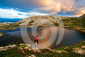 Man with red jacket celebrates with hands raised towards dramatic pink rain clouds and lakes outdoor in the wilderness in norway.