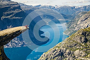 A man in a red jacket sitting on the Trolltunga rock with a blue lake 700 meters lower and interesting sky with clouds