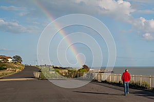 A man in a red jacket walks along a seaside promenade whilst a rainbow reaches down to the sea in front of him