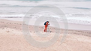 Man in red jumpsuit looking for lost luxuries with electronic metal detector on the beach on a cloudy fall day. He is