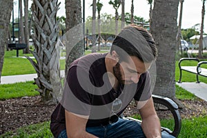 A man with a red shirt and blue jeans relaxes on a park bench playing with his aviator glasses looking down on a bright sunny day