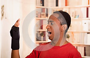 Man in red shirt staring at his own arm with shocked facial expression, wearing wrist brace support, blurry bookshelves