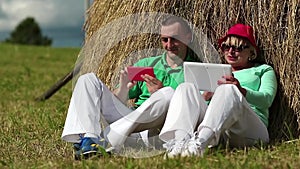 Man with red smartphone and woman with Tablet-PC sits near a haystack